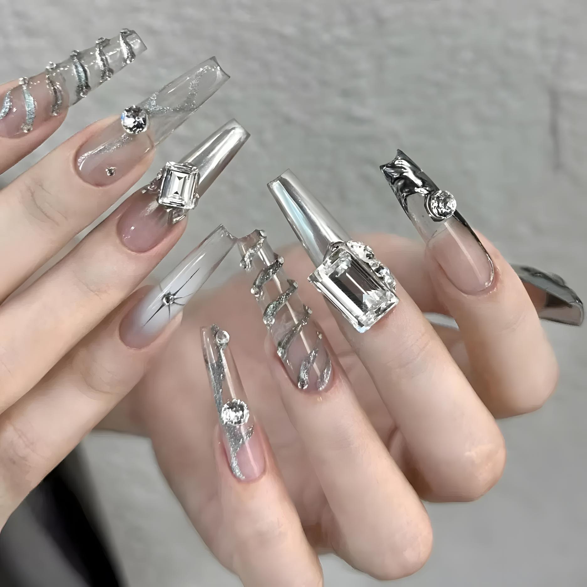 Close-up of a hand wearing MyLilith's extravagant press-on nails with a clear base and elaborate silver decorations, including rhinestones, geometric shapes, and metallic accents, in a long coffin shape.