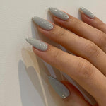 Close-up of a hand wearing MyLilith's grey press-on nails with a glossy finish, adorned with small rhinestone accents, in an almond shape.