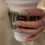 Close-up of a hand holding a coffee cup wearing MyLilith's grey press-on nails with a glossy finish, adorned with small rhinestone accents, in an almond shape.