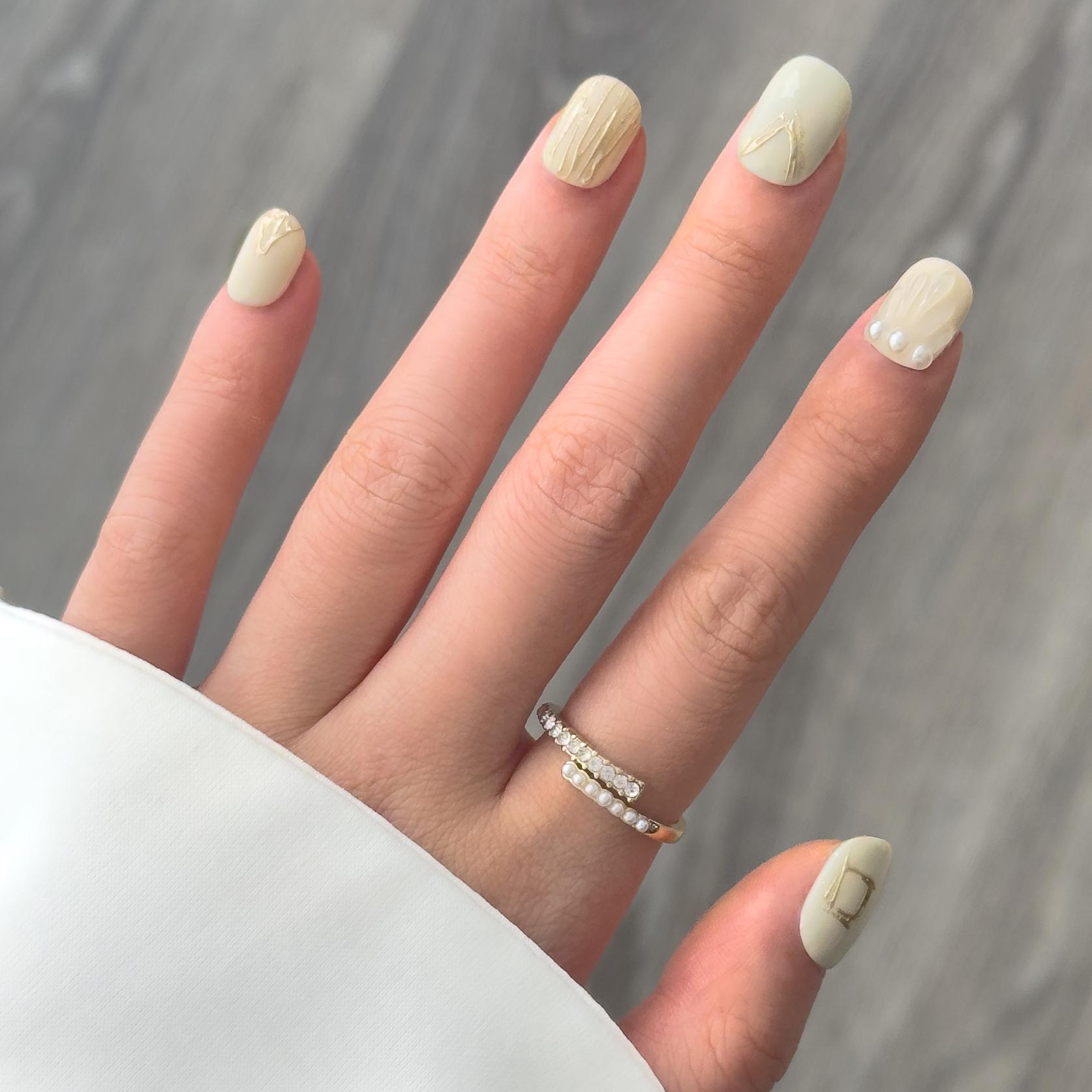 Close-up of a hand wearing MyLilith's press-on nails with a minimalist design, featuring matte beige nails with subtle gold accents and pearl details on one nail, in a short squoval shape.