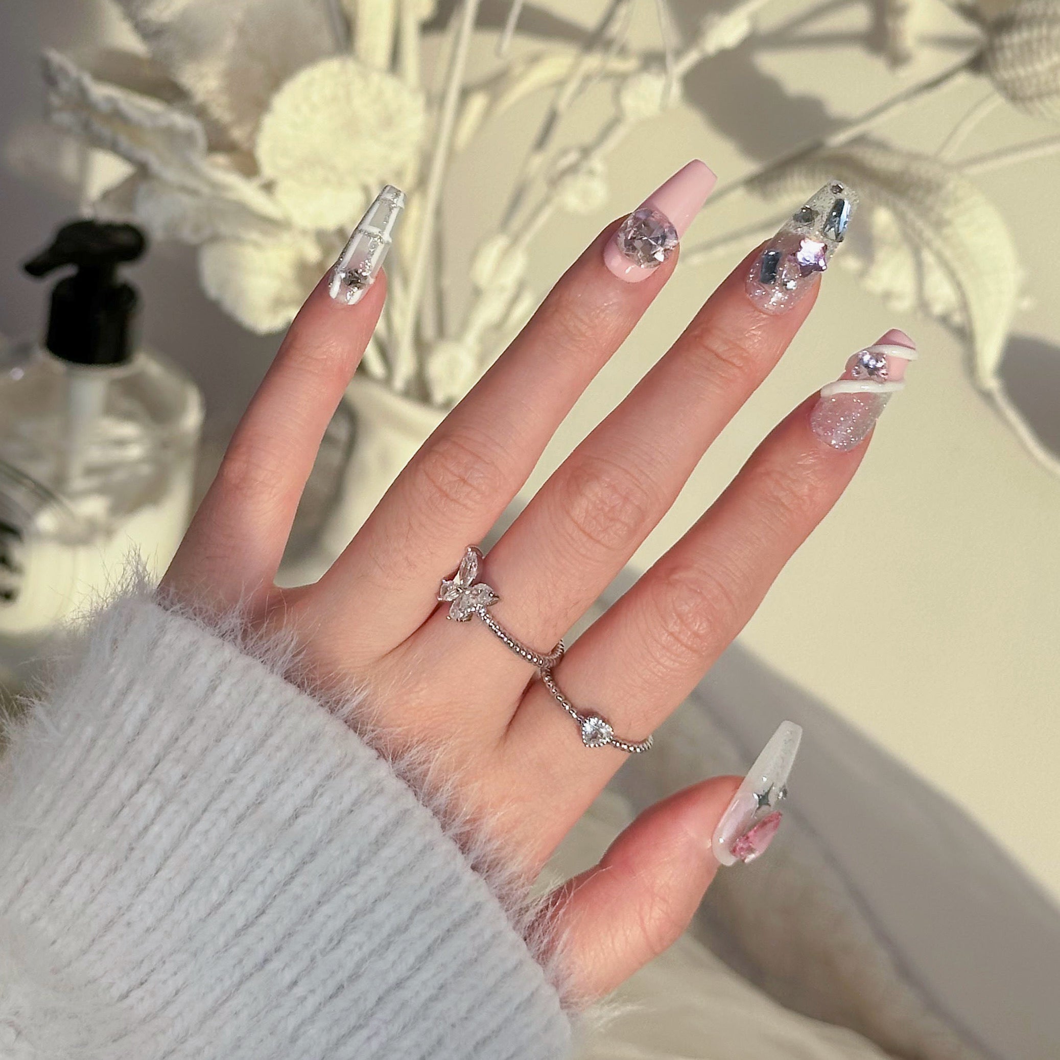 Back-view of a hand wearing MyLilith's press-on nails with a mix of designs, including clear, pink, and silver bases adorned with rhinestones, metallic accents, and intricate decorations, in a medium-length coffin shape.