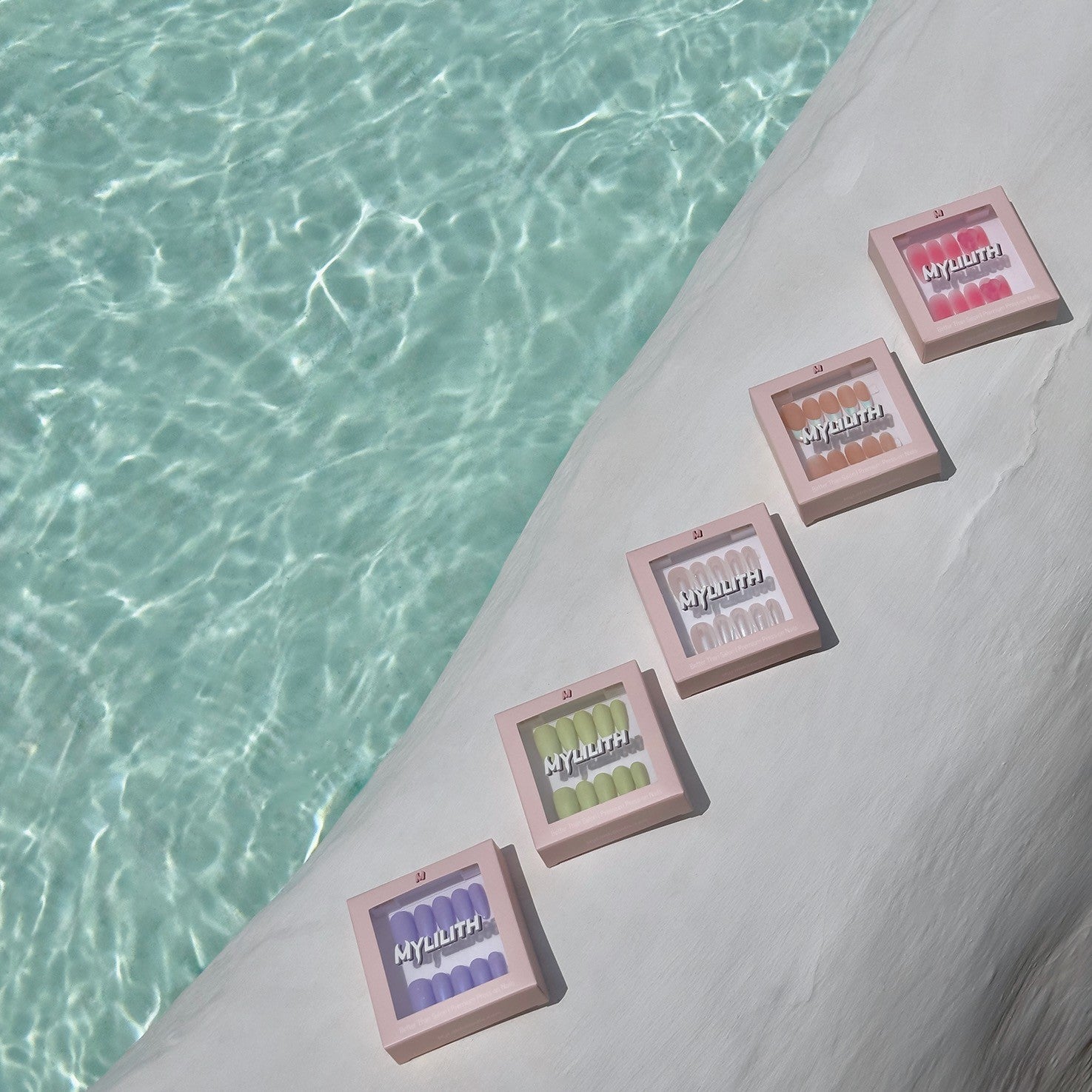 Five boxes of MyLilith Studio’s summer collection press-on nails displayed on a white surface beside a clear turquoise pool, showcasing different nail designs in pastel colours.