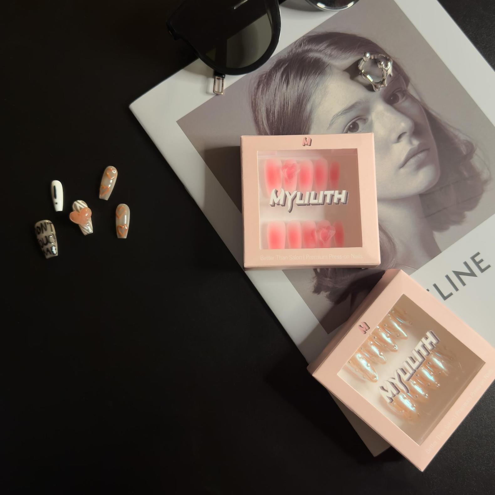 Two boxes of MyLilith press-on nails displayed on a dark surface alongside a fashion magazine, a pair of sunglasses, and several loose press-on nails.